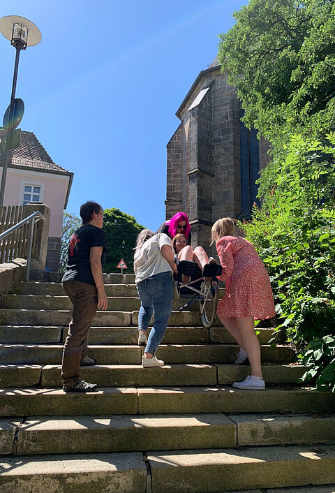 Schülerinnen der Fachschule Himmelkron tragen einen Rollstuhl die Treppe hoch.